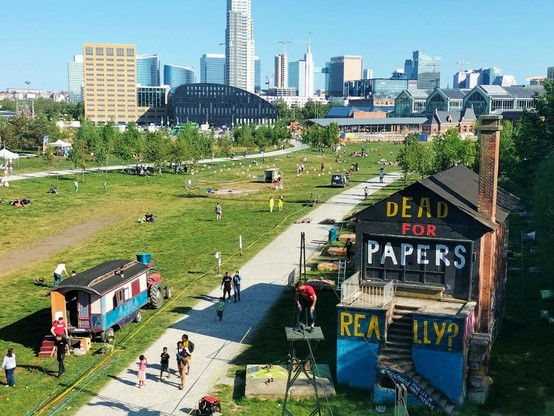 An urban park in Brussels. Several people are walking on the footpath or lounging on the grass. A colourful foodtruck and a small wooden building are visible in the foreground. The latter has the words "Dead for papers, really?" stenciled on in large letters. On the backdrop, the skyline of the Thur and Taxis quarter in north Brussels.