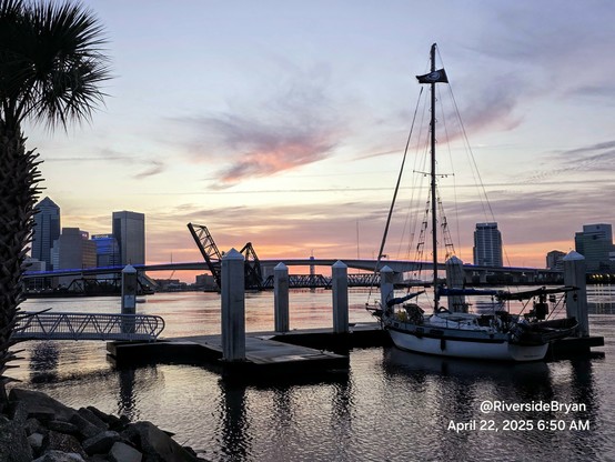 Early morning well before sunrise, overlooking a vast river and the downtown skyline with bridges. In the foreground a sailboat is tied up at a small marina. The city skyline and bridges are illuminated by pastel colors rising from the horizon in advance of sunrise.