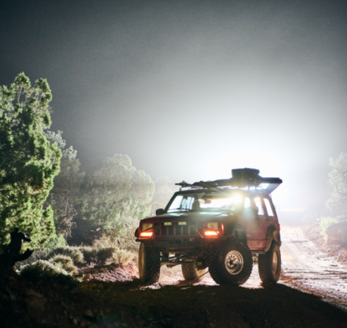 A jeep at night, with light shining from behind it. The front tires are both pointed inwards because the tie rod fell off.