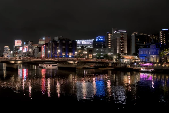 Night photograph of buildings overlooking a river in an entertainment distict. The bright white, yellow, red, blue, purple, lights emanating from the multi-story hotels and other nightlife buildings are reflected in the pitch black river water. The river is crossed by a bridge and several barges are docked around it.