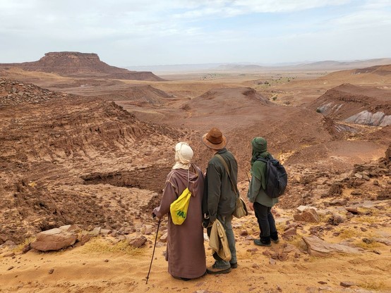 Landscape photograph of the rocky Sahara Desert in southern Morocco. There are three people standing in the foreground looking out over a dry valley.