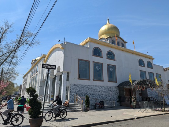A large gurdwara. It is a white building with a golden dome. Yellow flags can be seen. A middle aged woman is walking out, and two men on bicycles can be seen cycling past.