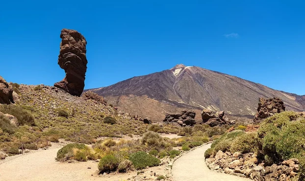 The image depicts a landscape featuring a prominent rock formation known as "Roque del Este" or "Roque del Teneguía" in the foreground, with a mountainous backdrop that includes the stratovolcano "Teide." This area is located in Teide National Park in Tenerife, Spain, and is characterized by its volcanic terrain, unique rock formations, and desert-like vegetation. The sky is clear and blue, contributing to the scenic beauty of the landscape.

Image Credits: Wikimedia / Thomas Wolf, www.foto-tw.de / CC BY-SA 3.0 de