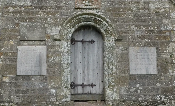 You see a grey old stone wall with lichens. In the middle a gray wooden door.