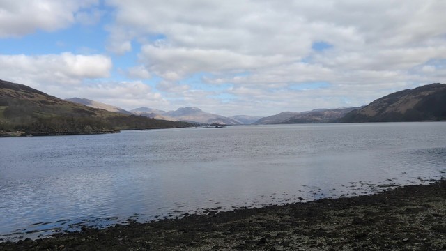 A loch with a rocky beach in the foreground and old mountains on the opposite side going back into the distance.