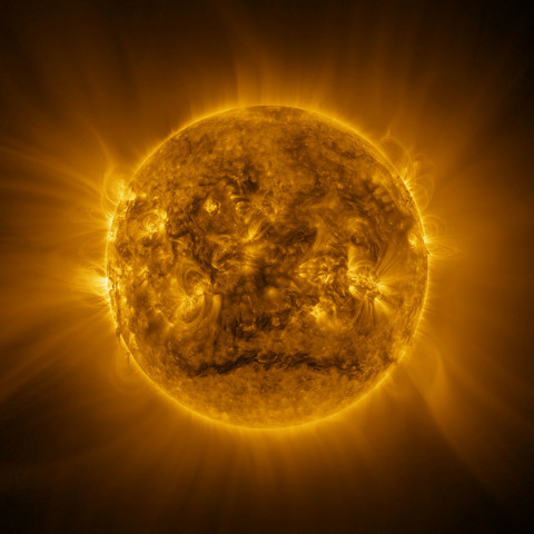 "The Sun looks like a warm yellow sphere with a surface covered with glowing messy hair. The yellow glow extends to the edges of the image, with some regions brighter than others. Many bright yellow arcs stick out from a wide band around the Sun’s equator. A darker region stands out across a roughly horizontal line near the Sun’s south pole. The bright arcs and some darker material can also be seen around the Sun’s edges."