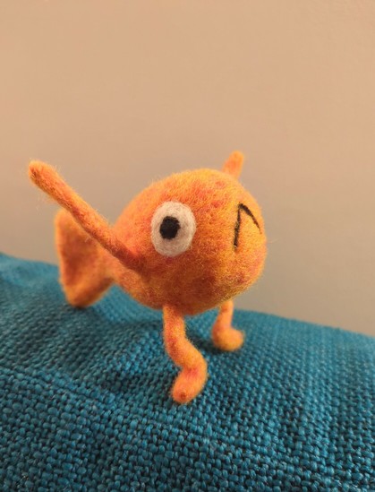 A small needle felted wool sculpture of Le Poisson Steve: an orange fish with arms, legs, big round eyes, and an inverted V for a mouth. Steve is standing on a blue couch cushion with legs bent and arms raised in the air.
