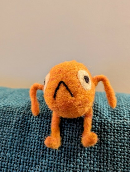 A small needle felted wool sculpture of Le Poisson Steve: an orange fish with arms, legs, big round eyes, and an inverted V for a mouth. Steve is sitting on a blue couch cushion, arms bent in a shrug and legs dangling over the edge.