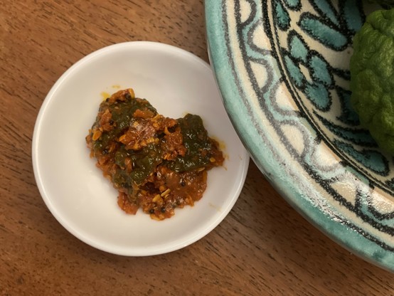 Some of the pickle in a small white bowl, next to a patterned bowl containing fresh makrut limes. The pickle has red spice mix encompassing the green lime pieces.