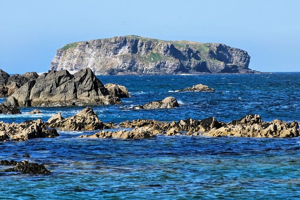A wide shot captures a rugged, rocky shoreline in the foreground with a larger, flat-topped island visible in the distance under a clear blue sky. The ocean water transitions from a lighter turquoise near the shore to a deeper blue further out.
