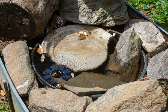 Six-inch diameter, rosette-shaped solar panel with a spray of water emerging from the center, inside a rock-filled bird bath.