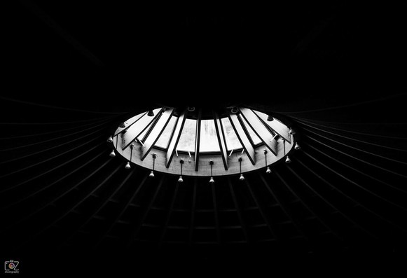 Low-key black and white photograph of a round skylight divided by wooden struts. The perspective from diagonally below makes the window appear elliptical. Numerous lamps hanging downwards are attached to the underside of the window structure. Struts of the ceiling can be faintly seen, which run out into the black background all around.
