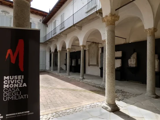 The image features an outdoor courtyard with arches and columns. There are historical stone artifacts displayed on black panels against the walls. To the left, there is a sign indicating the entrance to a museum, specifically mentioning "Musei Civici Monza" and "Casa degli Umiliati," suggesting it is a civic museum in Monza, Italy. The overall setting appears to be part of a museum or a cultural site.

Image Credits: Wikimedia / Musei Civici Monza / CC BY-SA 4.0