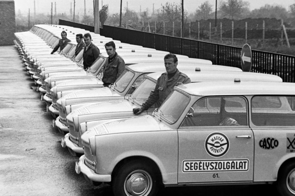 A monochrome photo of a long line of parked Trabant cars, all parked the same way. There must be at least thirty cars. On the side of each vehicle, there is the logo and words, in Hungarian, of the Hungarian Auto Club. There is in large letters on the driver's door the word 'Segélyszolgálat’, which translates in English to 'Emergency Services'. There are also some men standing between the vehicles at various points, looking towards the camera.