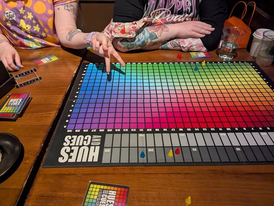 A game of Hues and Cues in progress. Two people's tattooed arms are visible on the other side of the table.