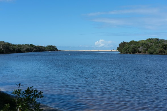 Photo of a wide creek looking towards the sandy mouth to the sea. The sky is blue.