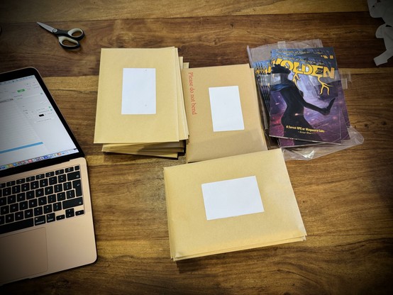 Several stacks of stuffed envelopes next to a stack of Wolden zines on a wooden table. A laptop with a spreadsheet and a pair of scissors surround it.
