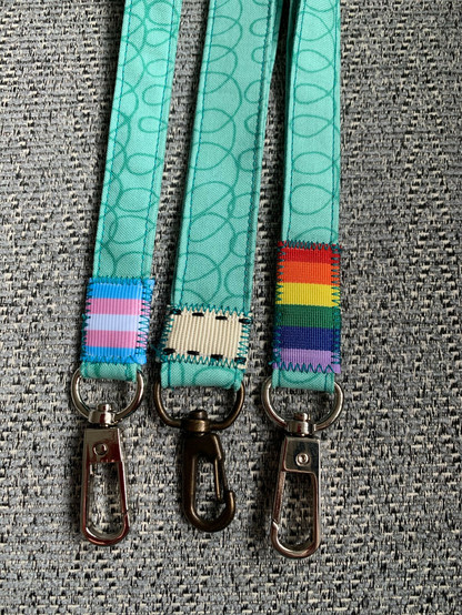 Three green cotton lanyards covered darker green swirls. At the end of one is a trans pride flag, then a blank ribbon, and finally a rainbow flag