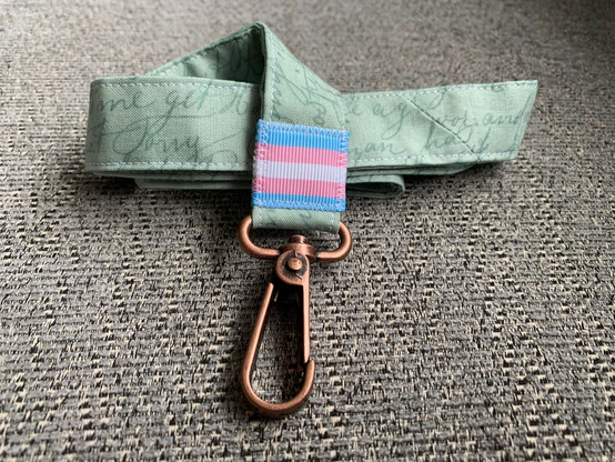 A green cotton lanyards covered in handwriting in a darker green colour. Near the lobster clip is a trans pride flag