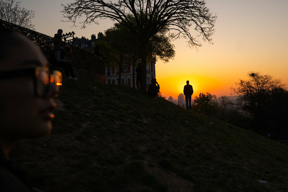 A man is watching the sunrise over the city of Paris. He stands directly  in front of the sun. in the background you can see the city, on the right some bushes. on the left part of a tree and houses and the face of a woman, she is wearing sunglasses. the light of the sun can be seen on the glass. 
the light looks golden because of the morning sun.