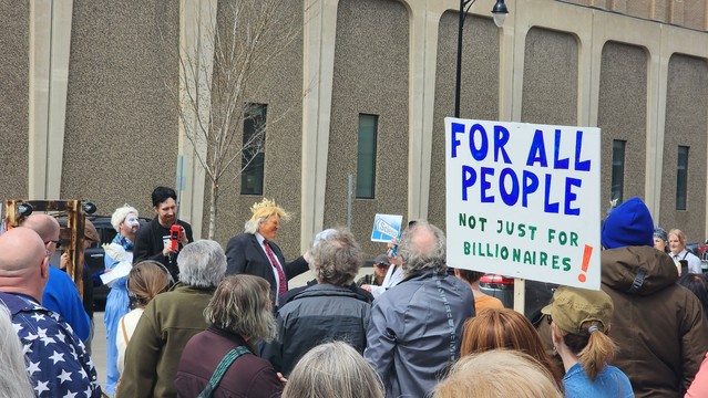 A crowd of people, a sign saya For All People Not Just For Millionaires. A Trump actor lookalike wears a gold crown.