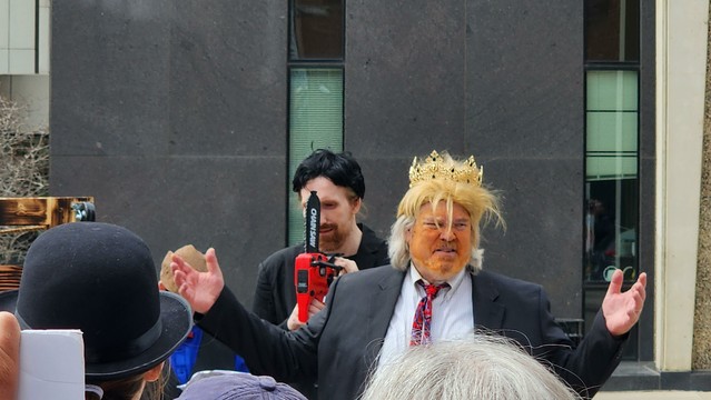 Close up of actor protestors dressed as Musk holding a chain saw, and Trump wearing a crown.