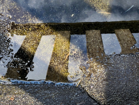 A puddle on a pebbled surface with a reflection of a wooden fence and a blue sky with clouds.