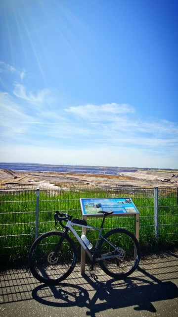 Ein Gravelbike ist angeleht an ein Hinweisschild, dahinter ein Gitterzaun und ein Streifen Wiese. Im Hintergrund das riesige Loch des Braunkohletagebaus Garzweiler. Der Himmel ist blau mit wenigen Wolken.