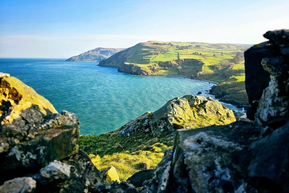 A scenic view of the Irish coastline with turquoise water meeting green, hilly land, framed by large, textured rocks in the foreground.