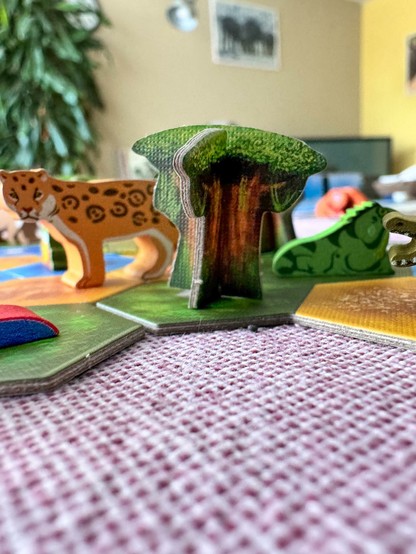 Das Bild zeigt eine Szene aus einem Spiel, die auf einem Tisch mit einem rosa und weißen gestreiften Tischdecken liegt. Im Vordergrund befindet sich ein Holzspielzeugbaum mit einem grünen Blätterdach und einem braunen Stamm. Neben dem Baum steht ein Holzspielzeugleopard mit orangefarbenem Fell und schwarzen Flecken. Im Hintergrund sind weitere Holzspielzeugtiere zu sehen, darunter eine grüne Echse Die Spielteile sind auf bunten, flachen Platten platziert, die verschiedene Farben und Formen haben. Im Hintergrund ist ein Teil eines Raumes mit einer Wand in Beige und einem Bild zu sehen.