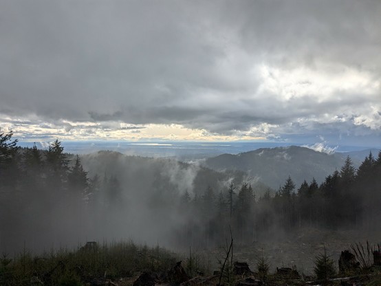 View from a mountain, with a cloudy sky. Lakes in the far background, trees in the middle ground.