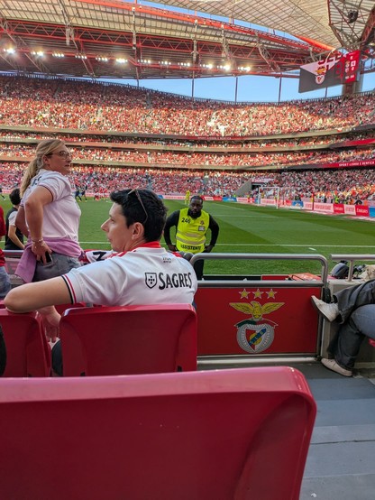 View of the interior of the Estádio da Luz during the game of SLBenfica against AVS Sad. The chair in front of me is empty, the rest of the place pretty crowded. Top right corner is the scoreboard, showing the halftime score of 4-0.