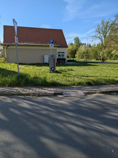 Auf einer Wiese etwa 4m vom Straßenrand steht eine AC-Ladesäule von N-ergie, mit Schild "P während des Ladevorganges". In der Wiese wächst Löwenzahn, keinerlei Befestigung des Untergrundes vor der Säule. Am Gehweg davor keine Absenkung, stattdessen nur ein paar grobe Hölzer, um das Befahren des Bordsteines zu vereinfachen.