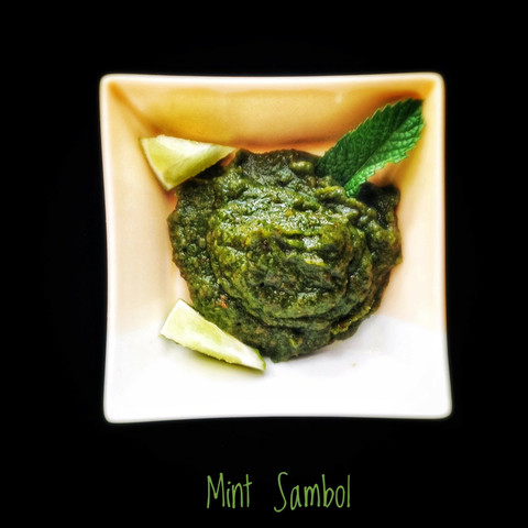 A green chutney in a square bowl, with a mint leaf and wedges of lime.