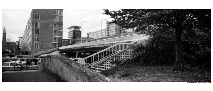 Black and white panorama photograph of a cross section of different stairs and railings of a bridge that crosses a canal. There are several apartment/office buildings in the back ground and a bike parked against a small motorboat cruiser.  Far left in the background is a church steeple and far right close up is a tree and undergrowth around some grass.