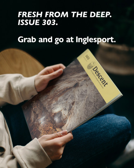 A person wearing a light beige sweatshirt and blue jeans is seated, holding a copy of Descent magazine, issue 303, which features a striking cave scene on its cover with rugged rock formations and two tiny cavers illuminated inside. The top of the image features bold white text that reads "Fresh from the deep. Issue 303. Grab and go at Inglesport." against a dark, blurred background of indoor plants and furniture.
