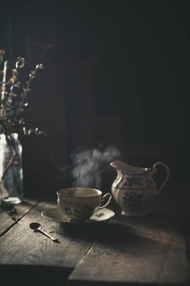 Alte Tasse, mit dampfendem Tee gefüllt. Ein Milchkännchen rechts daneben, links eine Glasvase mit trockenen Zweigen. Sonnenlicht fällt von rechts in das Bild.