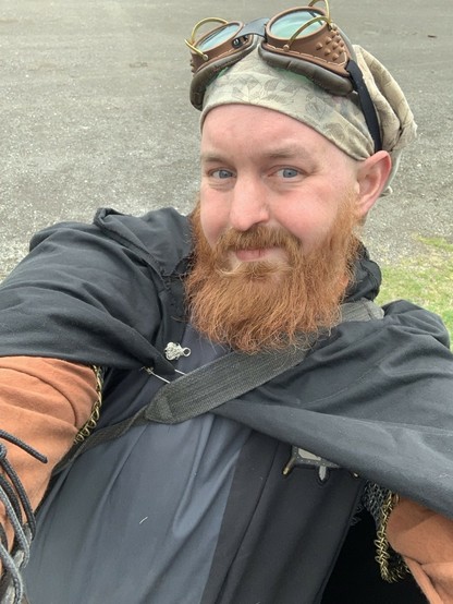 A cis white male with a red beard offers out a hug. He has a chainmail shirt underneath. He has a black and grey tabard, and a long sleeve brown shirt. He has googles on his head