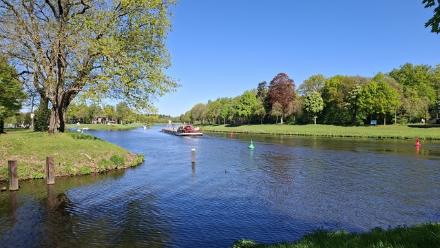 Foto der Ems. In den Fluss münden vorne und hinten jeweils Kanäle. In der Bildmitte befährt ein Binnenschiff das Wasser. Grüne und rote Bojen markieren die Fahrrinne. Am linken Bildrand steht ein großer Laubbaum am Ufer. Am gegenüberliegenden Ufer im Hintergrund der rechten Bildhälfte steht ein Wäldchen.