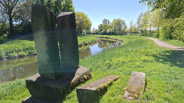 Foto der Vechte. Der Fluss fließt ruhig zwischen mit Gras bewachsenen Ufern. Im Hintergrund stehen beidseits des Flusses Bäume. In der rechten Bildhälfte führt ein Weg am Ufer entlang. Im Vordergrund steht eine Steinskulptur, bestehend aus zwei stehenden und drei liegenden großen Steinblöcken.
