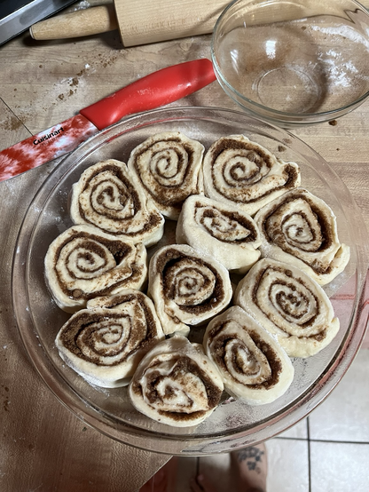 Photo of raw fresh cut cinnamon roll dough in a greased circular glass pie dish on a light wood countertop. A red tie dye knife in the background
