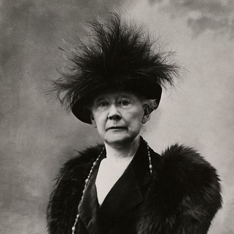 a photo of the artist, she is wearing furs and a large necklace, seen from her chest up, she is wearing a black hat with dark feathers sticking up out of it, she is looking at the viewer