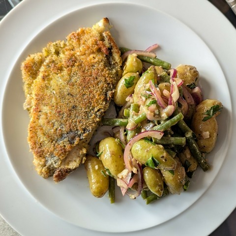 White plate, golden crumbed fillet of fish next to a pile of small potatoes, green beans and red onions all shiny with dressing