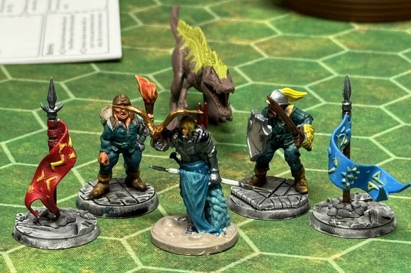 A group of painted miniature figures stands on a green hexagonal game board. The figures include a character with a torch, a knight in armor, and two other warriors holding weapons and flags, along with a small dinosaur-like creature in the background.