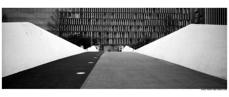 Black and white panorama photograph of a bike and pedestrian bridge taken from a low angle so the bridge takes up the whole bottom half of the photograph. In the background is a building with many black and white windows. The bridge is angular a white. The left half of the surface is pedestrian in dark black and the right side is for bikes, slightly lighter.