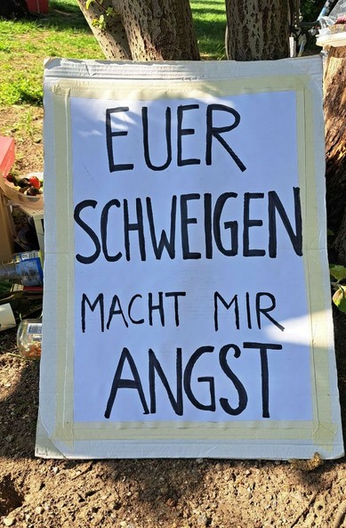 Plakat an Baum gelehnt, im Halbschatten mit schwarzer Schrift auf weissen Grund und einem Rahmen aus Kreppklebeband: "Euer Schweigen macht mir Angst"
