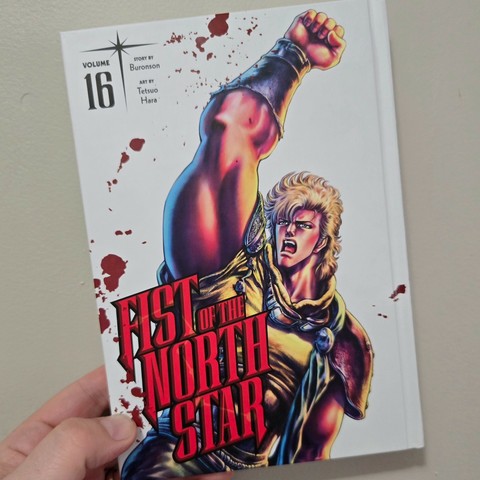 Photograph of a white person's hand holding Fist of the North Star, Volume 16, against a light colored wall. The hardcover book has a white cover with the number 16 in black text in the upper left-hand corner and "Fist of the North Star" in all red, uppercase lettering in the lower left-hand corner. A beefy, blonde man with his fist raised is on the cover behind the text. Blood streaks are strewn about the cover seemingly randomly.