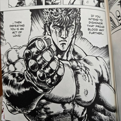 Photograph of a black and white manga panel from Fist of the North Star, Volume 16. Image shows Kenshiro (beefy man with fluffy dark hair and scars in the shape of stars on his pecs) standing shirtless with his fist outstretched in leather gloves and crying. He is saying, "If you intend to dishonor that proud blood any further...then defeating you is an act of love!"
