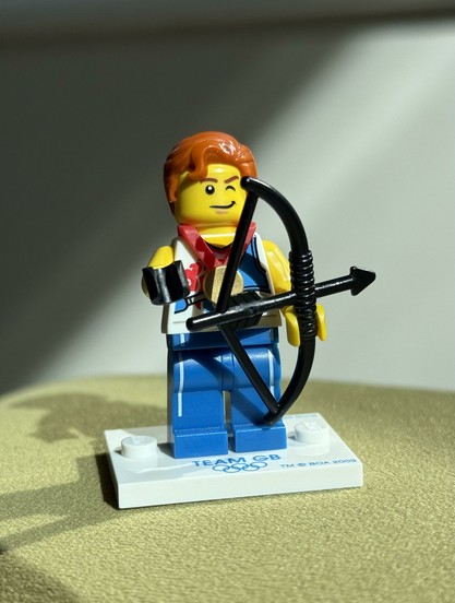 A LEGO mini figure representing an archer, featuring orange hair, a medal around its neck, and holding a bow and arrow. It stands on a white base labeled "TEAM GB". From the Olympic 2012 set.