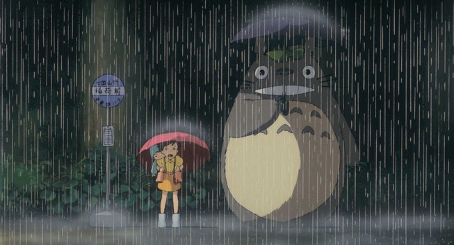 Image tirée de Mon Voisin Totoro.
On y voit l'image iconique de Totoro, une créature légendaire au sourire énorme qui attend à un arrêt de bus sous une pluie battante avec un parapluie, à ses côtés Satsuki qui porte sa petite soeur Mei sur son dos, également protégée par un parapluie.
